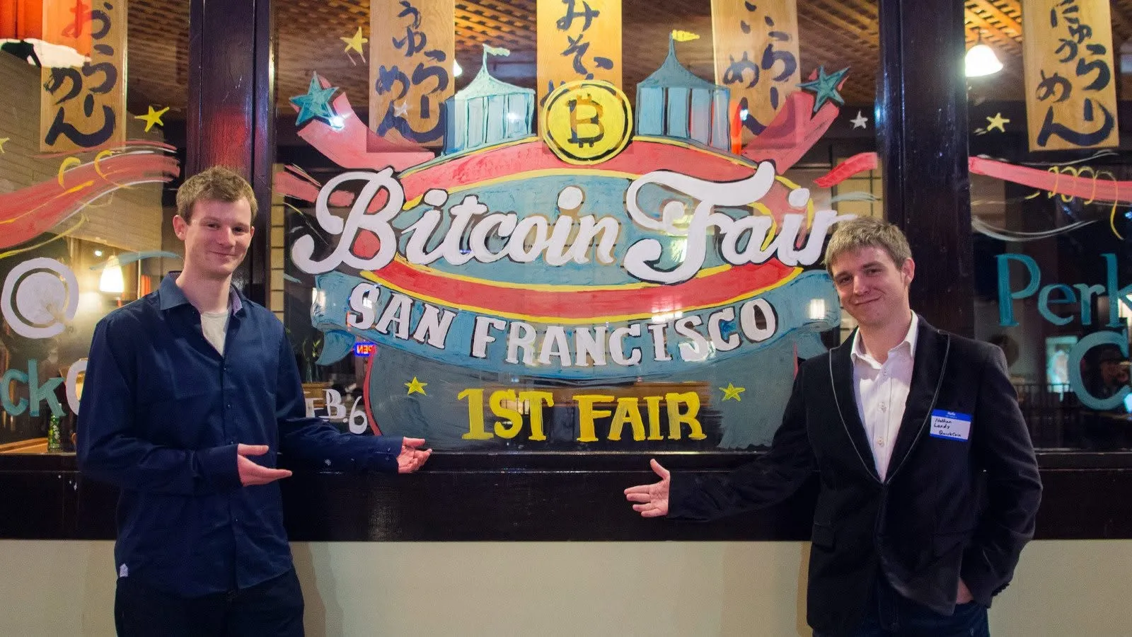 Two men gesturing toward a hand-painted window sign that reads Bitcoin Fair, San Francisco, 1st Fair