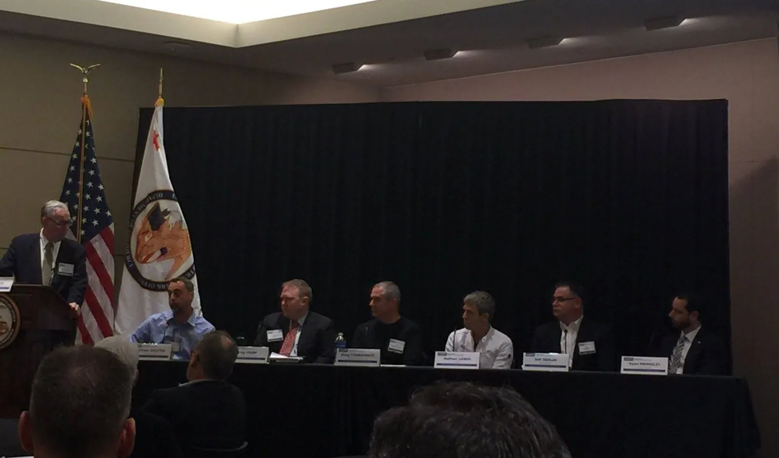 Six panellists seated at a long table beside a speaker at a lectern, an American flag behind them. One name card reads Nathan Lands.